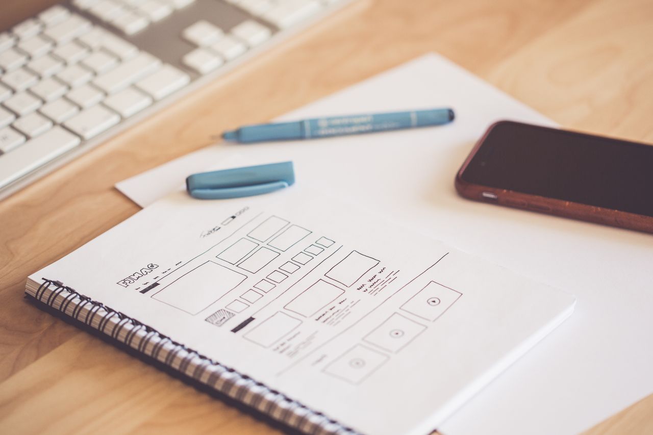 services-03 Un espace de travail soigné avec un carnet de croquis présentant des wireframes, un smartphone et un clavier sur un bureau en bois.